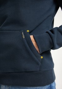 Mörkblå sweatshirt gjord av mjukt material, med en känguruficka, ribbade ärmslut och en gul accentknapp nära fickan.