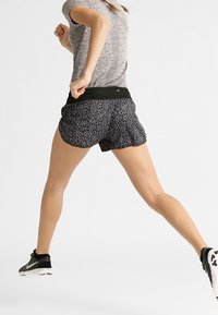 Pantalones cortos negros con un patrón geométrico, combinados con una camiseta deportiva gris, usados durante el movimiento. Presentan una cintura elástica y tela ligera.