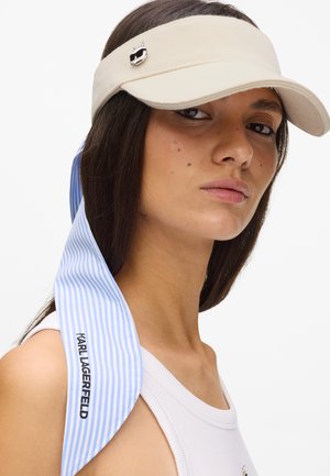 Femme portant une visière beige ornée d'une broche en forme de chat et un foulard noué à rayures bleues marqué "Karl Lagerfeld" sur de longs cheveux foncés, vêtue d'un débardeur blanc.