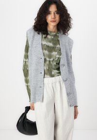 Grijze gebreide cardigan met knopen, gedragen over een groen tie-dye top. Lichtgekleurde corduroy broek en een zwarte pouch tas maken de look compleet.