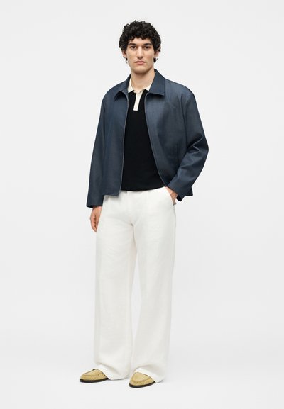 Νέος άνδρας που φορεί σκούρο μπλε μπουφάν, μαύρο-άσπρο πόλο, λευκό παντελόνι με φαρδύ πόδι και μπεζ παπούτσια, στέκεται μπροστά σε απλό φόντο.