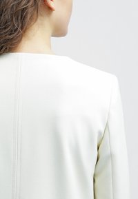 Vit skräddarsydd kavaj, strukturerad design, slät textur, minimala sömdetaljer och rakt skuren halsringning.