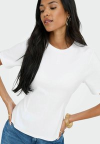 Vit kortärmad topp med rund halsringning, slät textur och subtila sidsömmar, parat med blå denimjeans och ett genomskinligt armband.