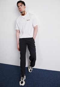 Hvid t-shirt med korte ærmer og Timberland-logo, parret med sorte bukser med sidelommer, samt sorte og cremefarvede sneakers med et unikt design.