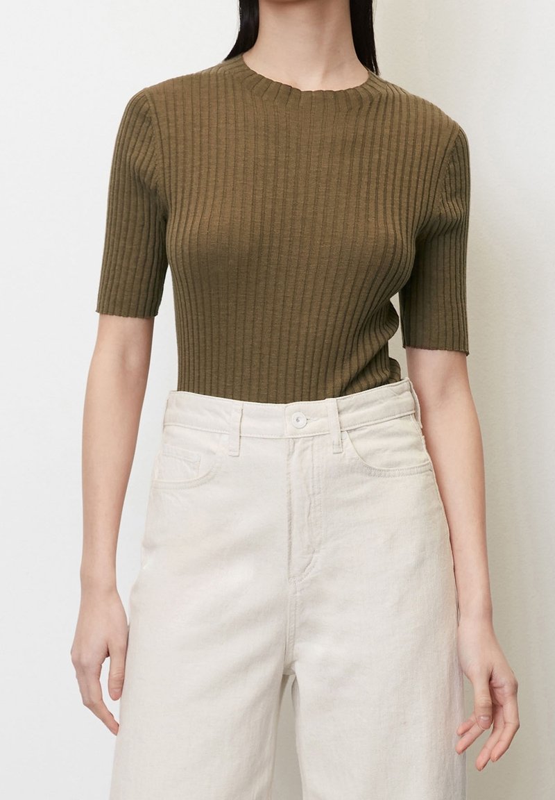 Personne portant un haut ajusté côtelé vert olive à manches courtes, associé à un pantalon large taille haute couleur écrue, sur un fond uni.