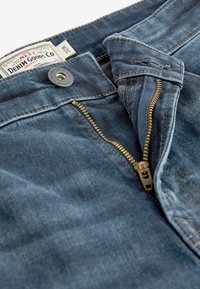 Tumedat sinised denim püksid, millel on nähtav kuldne tõmblukk, metallne nööp ja silt. Esitleb lähedalt vööosa ja esivedru.