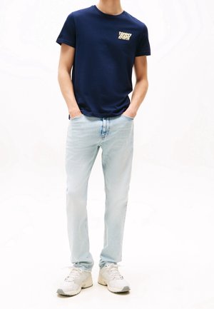 Mann trägt dunkelblaues Tommy Jeans T-Shirt, hellblaue Jeans, weiße Turnschuhe, steht mit den Händen in den Taschen vor weißem Hintergrund.