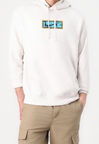 Vit huvtröja med en framficka, med en blå och svart grafisk design omgiven av en gul och svart leopardmönstrad kant.