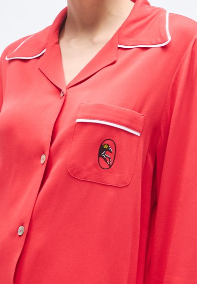 Červená košeľa na zapínanie s bielym lemovaním, jedným náprsným vreckom s vyšívaným motívom koňa, vyrobená z mäkkej tkaniny.