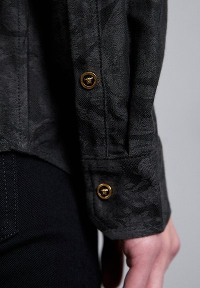 Manche en tissu texturé noir présentant un motif subtil, boutons dorés et bords soigneusement cousus, associée à un denim foncé.