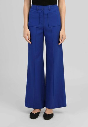 Pantalon bleu taille haute à jambes larges avec grandes poches plaquées devant, porté avec un top noir et des ballerines pointues noires sur fond gris.