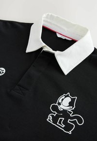 Camisa polo negra con un collar blanco, que presenta un diseño de un gato de dibujos animados en blanco en la parte frontal. Tejido suave con un ajuste estándar y mangas cortas.
