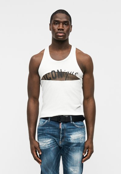 Άνδρας που φοράει λευκή αμάνικη μπλούζα με άνοιγμα και το κείμενο «ICONique», συνδυασμένη με σκισμένο μπλε τζιν και μαύρη ζώνη.