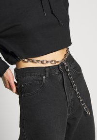 Persona che indossa jeans neri a vita alta e un maglione nero corto con una cintura a catena argentata avvolta attorno alla vita, pendente davanti.