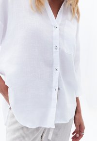 White linen button-up shirt with a v-neck, long sleeves, a single chest pocket, and a soft texture. Paired with striped light-colored pants.