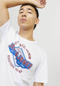 Witte t-shirt met korte mouwen, voorzien van een kleurrijk graphic logo met rode en blauwe accenten, gepresenteerd op een model met een neutrale achtergrond.