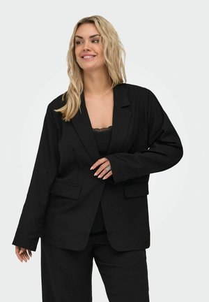 Usmievavá žena so svetlými vlasmi, oblečená v čiernom saku a čiernych nohaviciach na jednofarebnom svetlom pozadí.