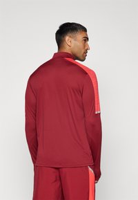 Mand set fra siden, iført en maroon langærmet sportjakke med lyse røde skulderpaneler og matchende shorts med røde sidestriber.