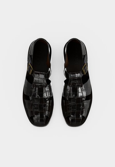 Μαύρα γυαλιστερά δερμάτινα ανδρικά σανδάλια με κλειστή μύτη, ανοιχτές πλευρές και κούμπωμα με πόρπη σε λευκό φόντο.