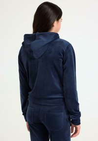 Survêtement en velours bleu marine, comprenant une veste à capuche et un pantalon ajusté avec poches arrière, texture lisse et tissu extensible.