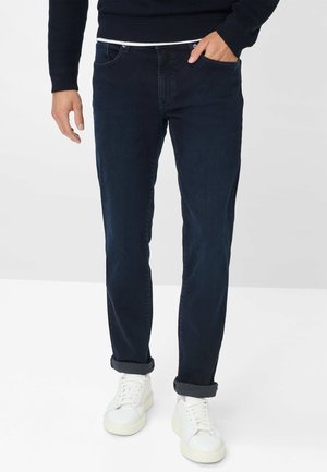 Dunkelblaue Jeans mit geradem Schnitt, umgeschlagenen Säumen, Five-Pocket-Design und getragen mit weißen Sneakers. Der Bund hat eine weiße Biese.