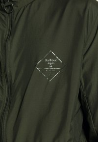 Vihreä kevyt takki, jossa on timantinmuotoinen logo ja heijastavalla hopeisella yksityiskohdalla olevat "Barbour"- ja "Beacon"-tekstit. Kankaan pinta on struktuuripintainen.