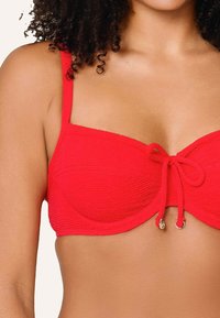 Top de bikini rojo texturizado con un diseño fruncido, tirantes ajustables y un lazo frontal con pequeños acentos de hardware dorados.