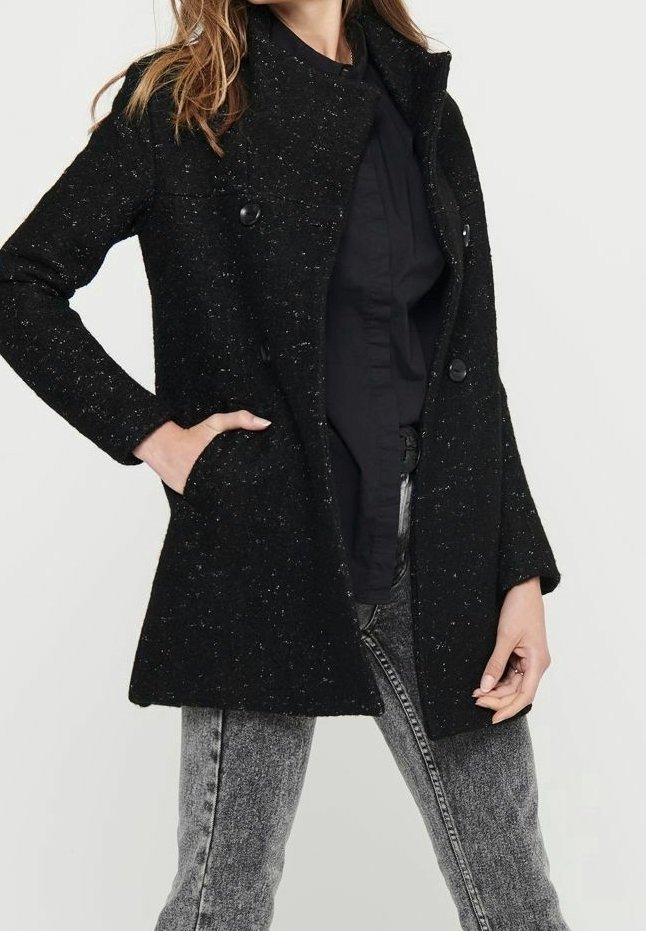 Manteau en mélange de laine noir avec une finition texturée, doté d'un col haut, de deux boutons et de poches latérales. Porté par-dessus une chemise noire et un jean gris.