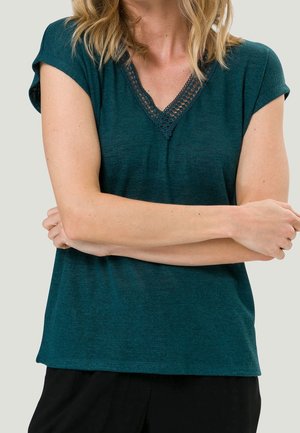 Kobieta o blond włosach, ubrana w turkusową bluzkę z krótkim rękawem i koronkowym dekoltem w kształcie litery V, z rękami skrzyżowanymi na tle gładkiego jasnego tła.