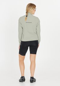 Lys grøn langærmet jakke med høj krave og reflekterende stripe på ryggen, parret med sorte cykelshorts og sorte cykelsko.