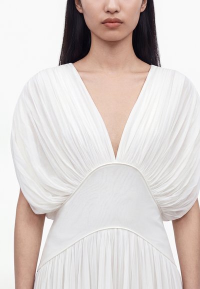 Γυναίκα που φοράει λευκό φόρεμα με βαθύ ντεκολτέ σε σχήμα V και φουσκωτά, πιετά κοντά μανίκια, εμφανιζόμενη από τους ώμους μέχρι τη μέση.