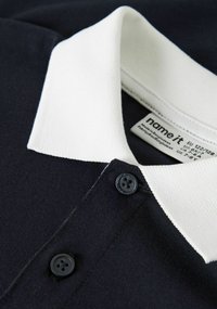 Polo azul marino con un cuello acanalado blanco, que presenta dos botones negros y una etiqueta que indica la talla y las instrucciones de cuidado.