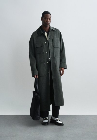 Vīrietis ģērbies garā tumši zaļā mētelī, melnās biksēs, baltos un melnos mokasīnēs, turot melnu somu, stāv uz vienkārša gaiši pelēka fona.