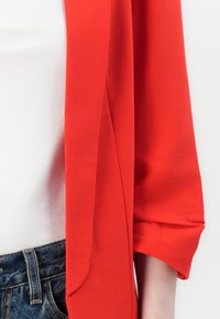 Jasnoczerwony, lekki żakiet w stylu kimona o luźnym kroju i podwiniętych rękawach, zestawiony z białą bluzką i dżinsowym pasem.