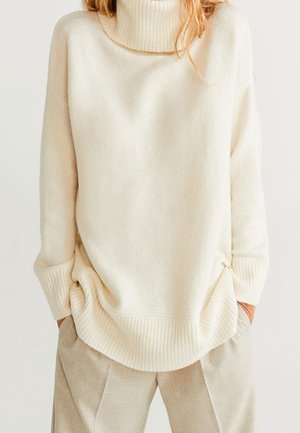 Femme portant un pull à col roulé ample crème et un pantalon beige clair, les mains dans les poches, sur un fond uni.