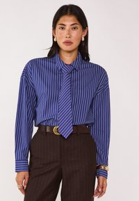 Camicia a righe blu con maniche lunghe e cravatta, indossata con pantaloni marroni a righe sottili. I dettagli includono un colletto e maniche con polsini.