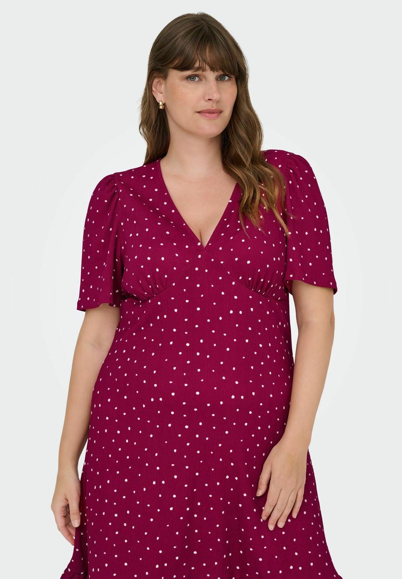 Bordo haljina s bijelim točkicama, kratkim napućenim rukavima i V-izrezom. Tkanina ima teksturirani uzorak i lepršavu siluetu.