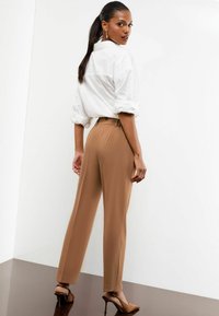 Braune weite Hosen aus glattem Stoff, kombiniert mit einer weißen Bluse. Das Model steht vor einem hellen Hintergrund und zeigt die Passform des Outfits.