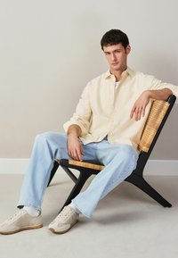 Jonge man zittend op een zwarte rattanstoel, gekleed in een lichtgeel overhemd, lichtblauwe jeans, witte sokken en beige sneakers, tegen een grijze muur.