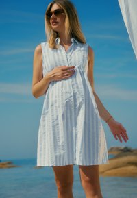 Lichtblauwe en witte gestreepte mouwloze jurk met een knoopsluiting aan de voorkant, losse pasvorm en zakdetails. Buiten gedragen tegen een blauwe lucht.