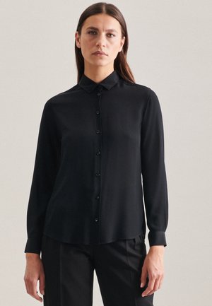 Maglietta nera a maniche lunghe con colletto, chiusura frontale a bottoni e tessuto liscio. Polsini affilati e vestibilità sartoriale visibile.