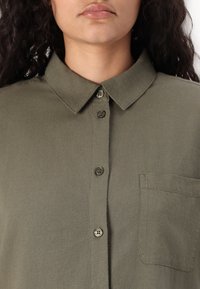 Camisa verde-azeitona de botão, feita de tecido macio, com colarinho, quatro botões e um bolso no peito com um acabamento simples e texturizado.