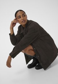 Brun ruskindsjakke med krave, trykknapper og lange ærmer. Model i squat, iført sorte støvler. Minimalistisk baggrund.