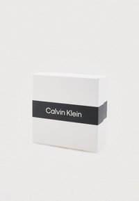 Boîte rectangulaire blanche avec une bande noire portant le texte "Calvin Klein" à l'avant. Le design est minimaliste et épuré.