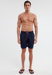 Tamsiai mėlynos maudymosi šortai su elastinga juosta ir raišteliais, turintys šonines kišenes ir minimalistinį dizainą. Dėvimi su juodais šlepetėm.