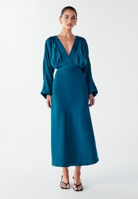 Žena v tmavě modré saténové dlouhé šaty s hlubokým V výstřihem, stojící na pozadí bílého pozadí, obutá v černých otevřených lodičkách.