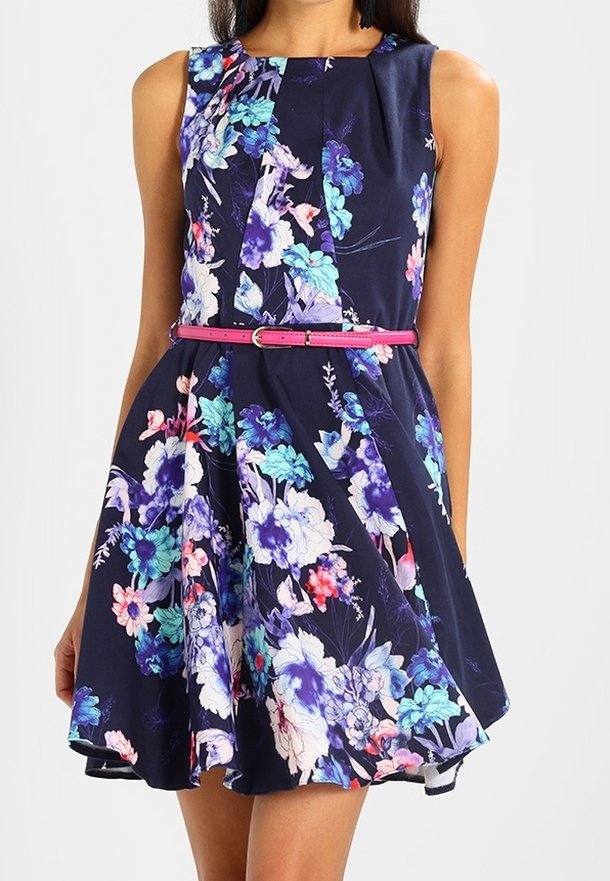 Femme portant une robe bleu marine sans manches avec des motifs floraux colorés et une fine ceinture rose à la taille.