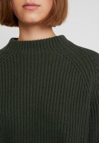 Frau trägt einen dunkelgrünen, gerippten Strickpullover mit hohem Rundhalsausschnitt und kurzem braunem Bob-Haarschnitt.