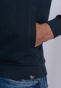 Sudadera azul marino con un bolsillo canguro, puños y dobladillo acanalados. Presenta una etiqueta negra con el texto "Petrol" en la parte inferior.