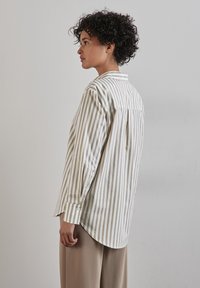 Camicia di cotone con righe verticali beige e bianche, dotata di colletto, maniche lunghe e orlo curvo, indossata sopra pantaloni beige chiari.